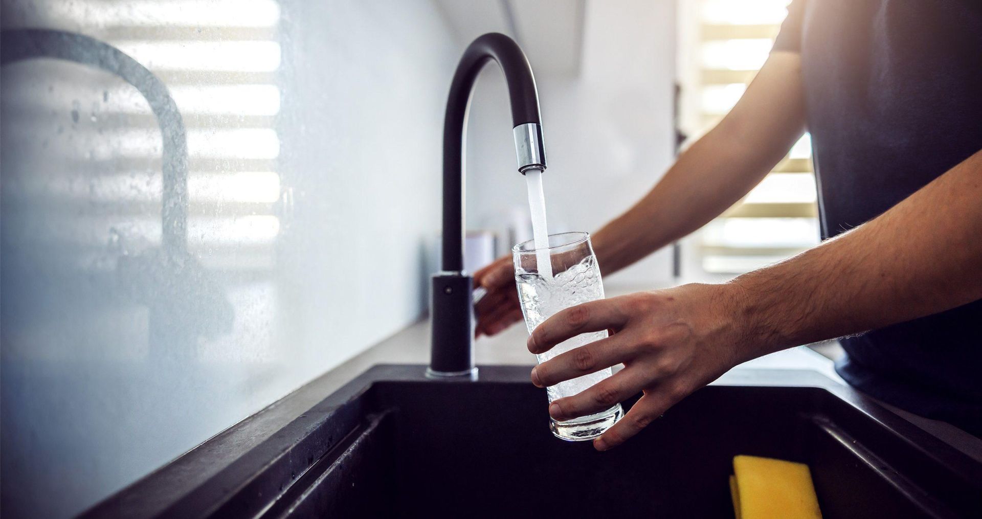 Male arms filling a clear glass with water at a clean, modern kitchen sink.