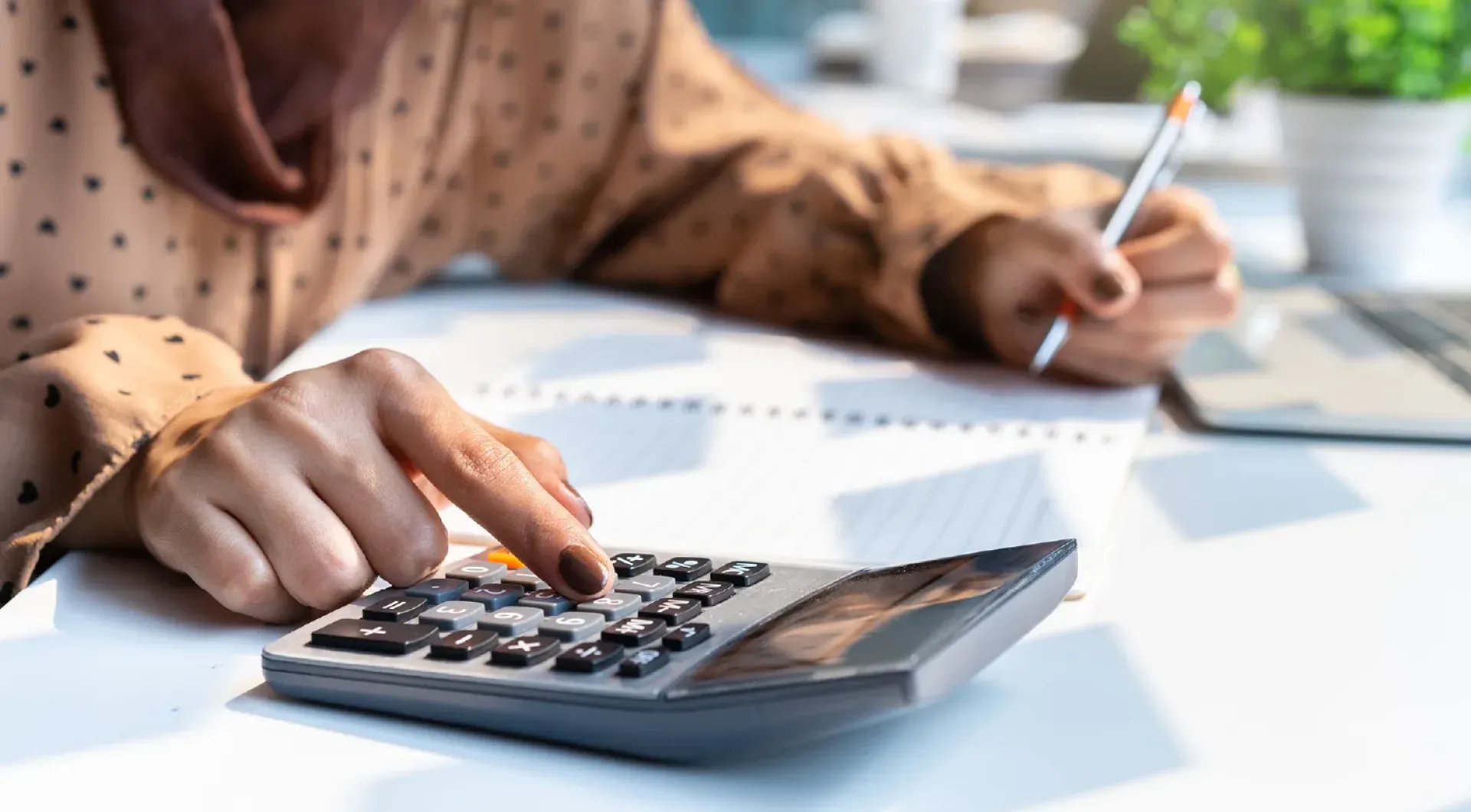 Hands and torso of female at desk, using a calculator and writing in a notebook with laptop nearby.