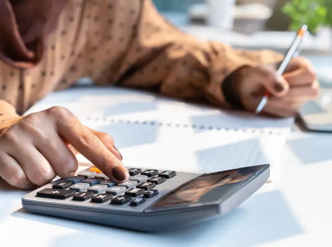 Hands and torso of female at desk, using a calculator and writing in a notebook with laptop nearby.