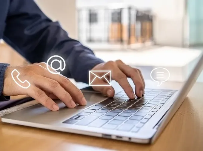 Hands of person wearing blue button-down shirt, typing on a silver laptop on a table in well-lit room.