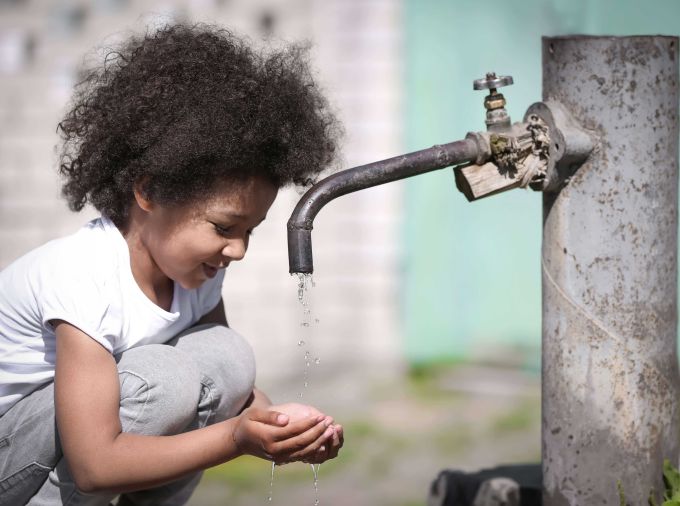 Girl squats with hands together to collect water from an outdoor spigot.