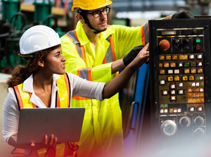 Two engineers in yellow vests and construction helmets examine equipment while one holds a laptop.